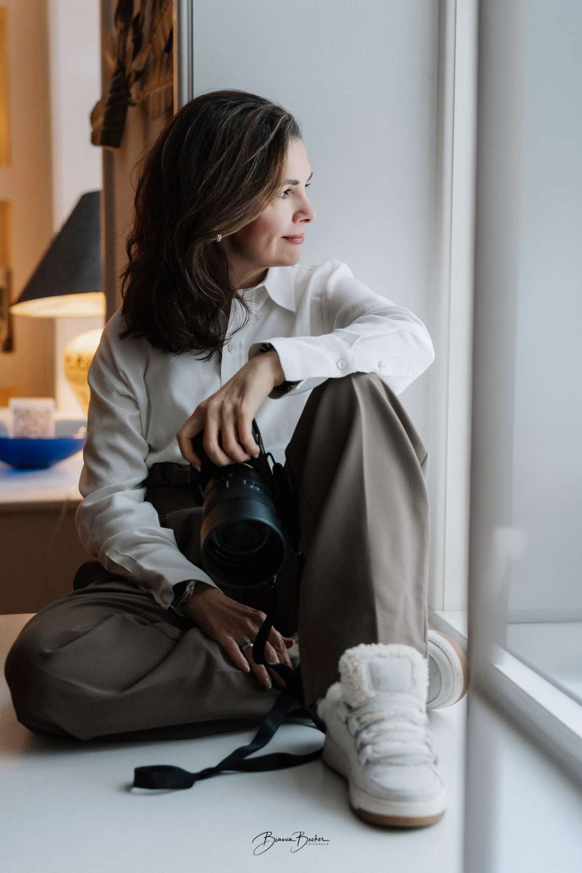 Porträtaufnahme von Bianca Becker, Friseurmeisterin und Make-up Artistin in ihrem Studio, lächelnd mit Kamera in der Hand.